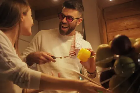 Warm lifestyle scene of a smiling man and woman cooking together in a wooden interior; natural gestures, food preparation and amber light convey hospitality, connection…