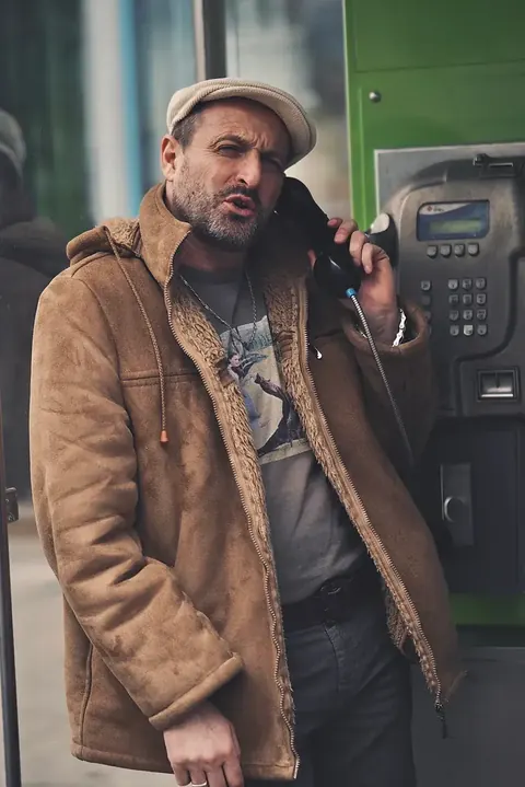 Street character portrait of a bearded man in a cap and layered brown clothing using a public telephone; muted tones and weathered textures build an authentic urban…