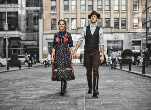 Full-length couple portrait in coordinated vintage-inspired clothing on a European square; their joined hands and measured stride create a coherent shared identity.