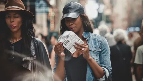 Street-documentary scene of two women reading and sharing printed material in a crowded city environment; gesture and attention communicate curiosity, culture and social…