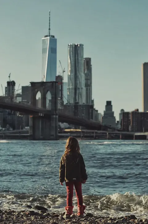Small figure facing the Manhattan skyline across the water; the monumental city scale turns the scene into a visual statement about aspiration, distance and possibility.
