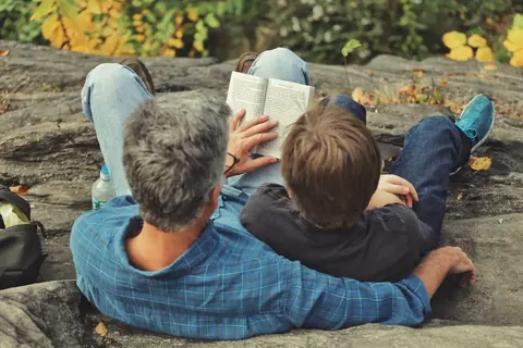 Overhead family scene of an adult and child lying together outdoors while reading; the close framing and autumn setting emphasise safety, attention and intergenerational…