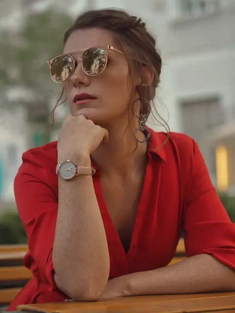 Close editorial portrait of a woman in a red dress and sunglasses resting her chin on her hand; the restrained gesture and accessories suggest composed, cosmopolitan…