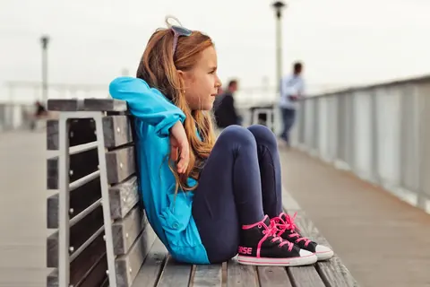 Youth lifestyle portrait of a girl seated beside a waterfront railing in bright casual clothing; folded posture and distant gaze create an introspective urban-travel…