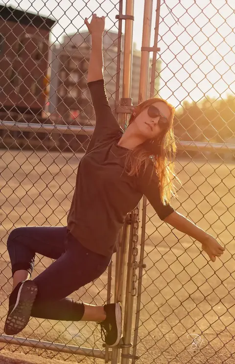 Urban fashion portrait of a woman leaning against a chain-link fence in late sunlight; angled posture and industrial texture create a rebellious, contemporary identity.