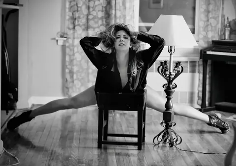 Black-and-white interior portrait of a woman seated backwards on a chair beside a lamp; symmetrical posture and direct gaze create a strong, theatrical character study.