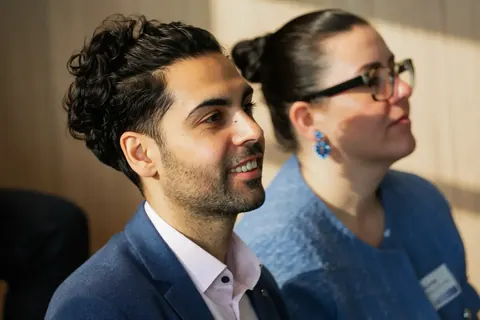 Candid audience reaction at a professional gathering, with two guests smiling and listening; the image records engagement, atmosphere and human connection.