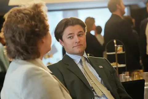 Candid conversation at a business reception between a seated man in a suit and a woman seen from behind; facial attention and surrounding guests convey active networking.