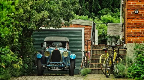 Vintage automobile and bicycle parked beside a small garden garage; weathered objects and dense greenery create a quiet study of time, memory and mechanical character.