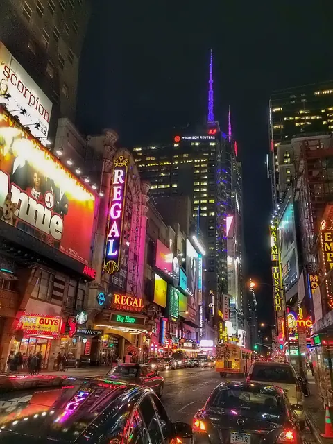 Night view of Times Square filled with neon signs, traffic and wet reflections; compressed urban light conveys spectacle, density and sleepless metropolitan energy.
