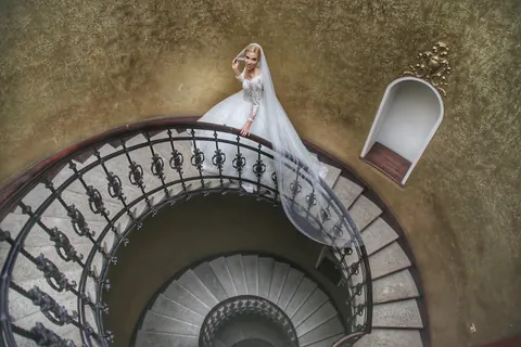 Bride in a white gown standing at the top of a circular staircase; the sweeping geometry and elevated viewpoint transform the scene into a formal study of anticipation…
