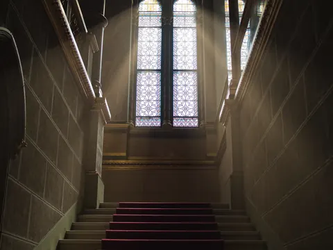 Symmetrical stone stairwell rising toward a tall stained-glass window; converging lines, red carpet and cool daylight create ceremonial depth and architectural silence.