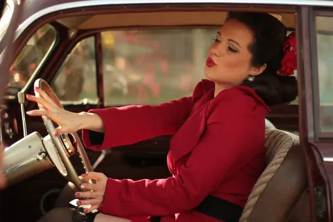 Vintage-styled woman in a red outfit seated at the wheel of a classic car; gesture, colour and interior detail build a cinematic story of elegance and control.