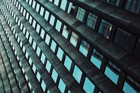 Upward view of a modern building façade with repeating dark windows and turquoise reflections; converging geometry turns architecture into an abstract rhythm.