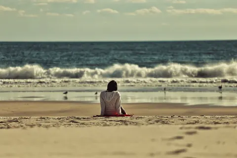 Solitary woman seated on a broad beach facing the sea; waves, open horizon and small human scale create a contemplative image of distance and inward attention.