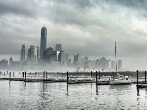 Manhattan skyline emerging through fog across the water; muted tones, empty docks and softened towers convey uncertainty, memory and atmospheric scale.