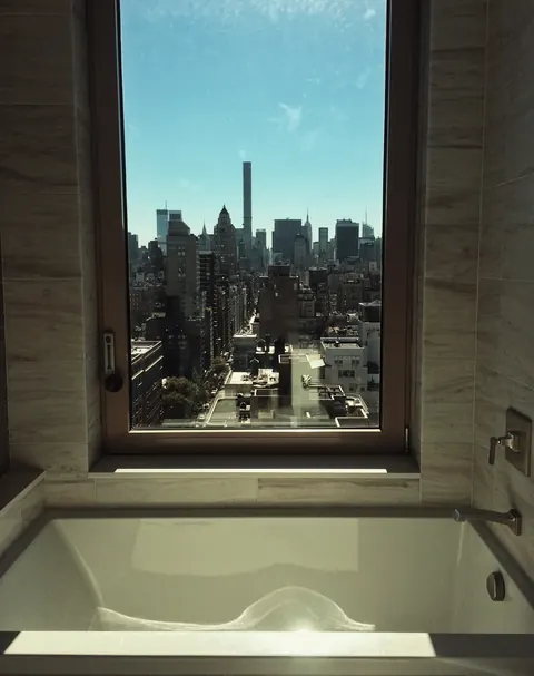City skyline framed through a narrow bathroom window above a white bath; the intimate interior and distant towers create a dialogue between privacy and metropolis.