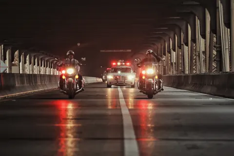 Police motorcycles escorting a vehicle through a dark bridge at night; wet reflections, symmetry and warning lights create cinematic movement and institutional power.