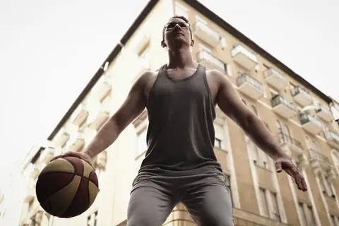 Low-angle urban portrait of a basketball player holding a ball between tall residential buildings; body scale and architecture turn athletic readiness into a study of…