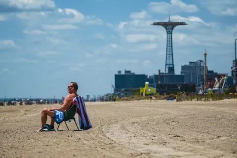 Solitary figure seated on a broad beach with the Coney Island Parachute Jump in the distance; large negative space evokes memory, waiting and the human scale within…