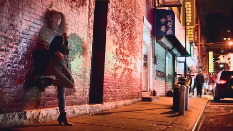 Night street portrait of a woman leaning against a red-lit brick wall; mixed urban light and distance create a cinematic narrative of solitude and self-protection.