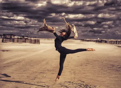 Dance photograph on an empty beach beneath dramatic clouds; extended limbs and windblown fabric translate technical movement into freedom and emotional release.
