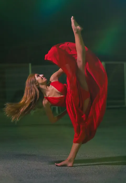 Performance portrait of a dancer in flowing red fabric executing a high extension against a dark coloured background; motion and costume create dramatic visual energy.