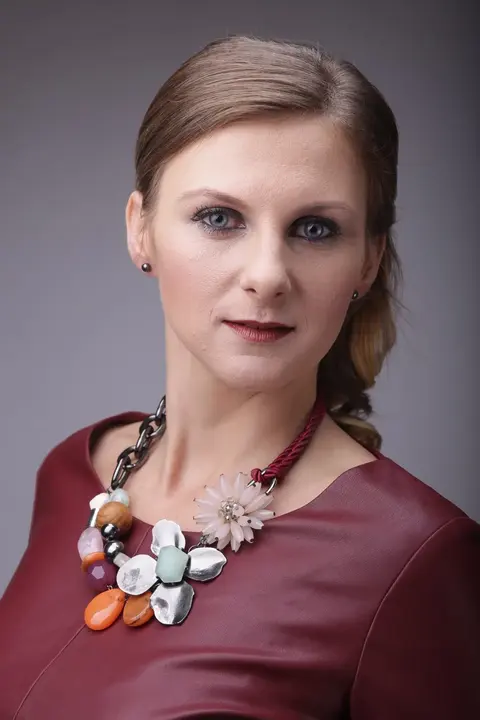 Formal studio headshot of a woman with carefully styled hair, burgundy clothing and a statement necklace; neutral light keeps attention on expression, competence and…
