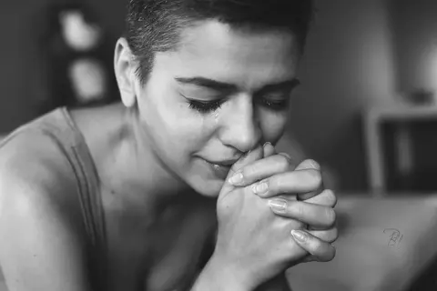 Intimate black-and-white portrait of a woman with closed eyes and hands clasped near her face; close framing and tonal restraint suggest reflection, vulnerability and…