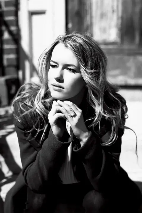 Black-and-white editorial portrait of a seated woman in a dark blazer, hands gathered beneath her chin; window light and thoughtful posture create intellectual calm.