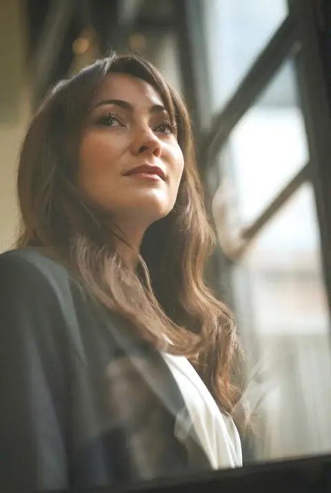 Environmental portrait of a woman in a bright interior looking upward toward window light; layered reflections and architectural lines give the image aspiration and quiet…