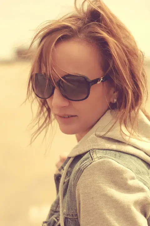 Sunlit lifestyle portrait of a woman in sunglasses and a light jacket looking down over her shoulder; warm colour and loose hair create relaxed, contemporary elegance.