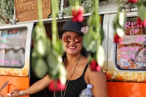 Colourful environmental portrait of a smiling woman at an urban food or market stand, partially framed by hanging flowers; the scene communicates openness and local…