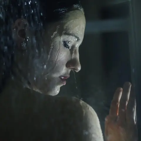 Moody profile portrait of a woman seen through rain-streaked glass; cool colour, reflection and a raised hand produce a cinematic sense of distance and introspection.