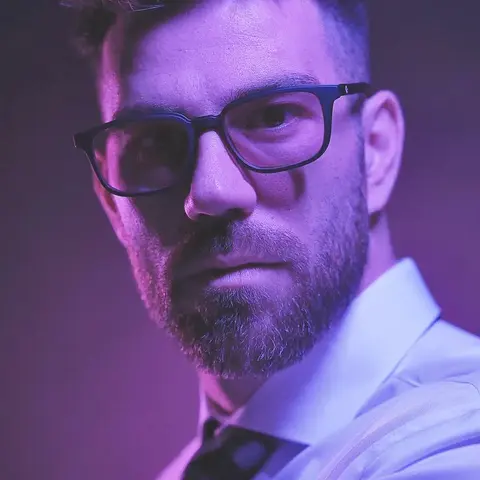 Low-key portrait of a bearded man in glasses and formal clothing under purple light; colour contrast and close framing create a contemporary executive or creative-leader…