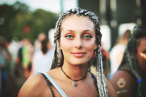 Street portrait of a woman with braided hair and vivid eye makeup at a public event; shallow depth of field isolates a confident, individual style.