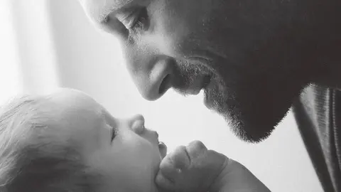 Tender black-and-white close-up of an adult man touching his face to a baby’s forehead; minimal framing centres trust, protection and parental intimacy.