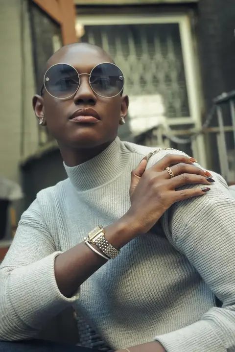 Urban fashion portrait of a woman in round sunglasses and a light knit top, hands gathered near her shoulder; low-angle framing and architectural background communicate…