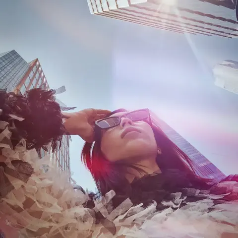 Low-angle city portrait of a woman in sunglasses framed by tall buildings and bright sun flare; the perspective expresses ambition, scale and metropolitan identity.