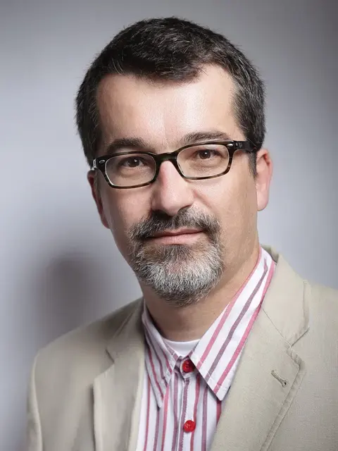 Professional studio headshot of a man with glasses, moustache and beard in a light jacket; neutral background and measured expression communicate expertise and trust.