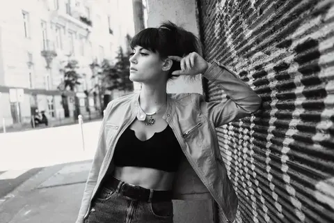 Black-and-white street-fashion portrait of a woman leaning beside a textured urban wall; cropped clothing, jewellery and closed eyes emphasise self-expression and…