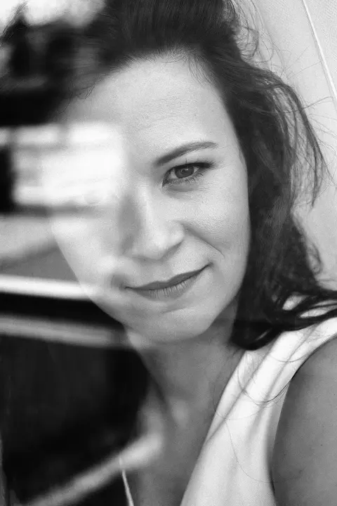 Layered portrait of a woman seen through glass or reflection, with one eye sharply visible; the fragmented foreground suggests observation, privacy and complex identity.