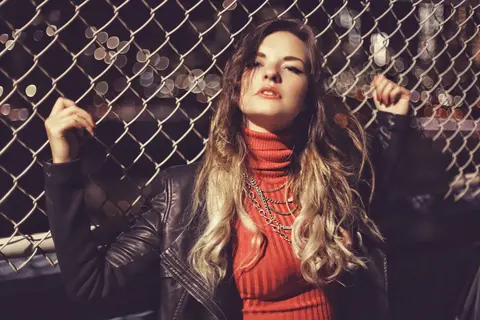 Fashion portrait of a woman in a red turtleneck and leather jacket posing against a chain-link fence; strong posture and colour create a bold metropolitan persona.