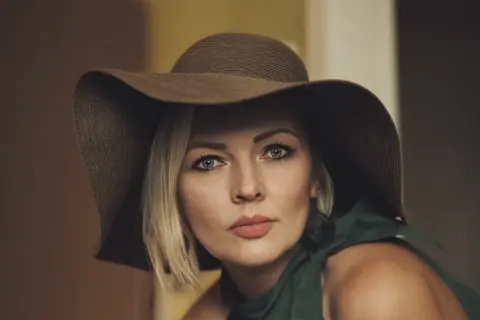 Close editorial portrait of a woman wearing a broad dark hat; direct blue-eyed gaze, shallow focus and restrained colour create classic cinematic elegance.