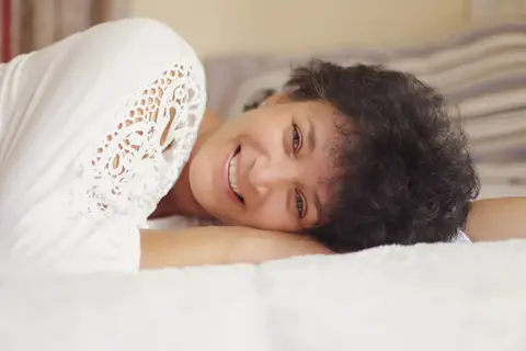 High-key lifestyle portrait of a smiling woman with short curly hair lying on a bed; white textures and relaxed posture communicate comfort, openness and joy.