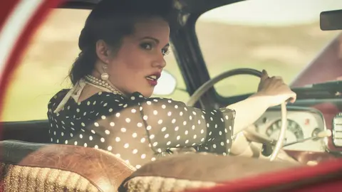 Vintage-inspired portrait of a woman in a polka-dot dress and gloves seated at the wheel of a classic car; styling and warm colour build a narrative of glamour and…