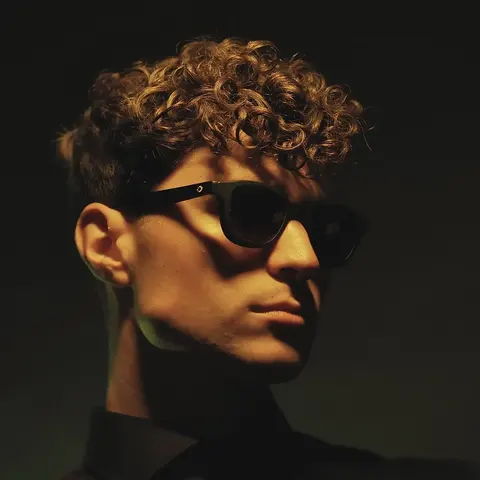 Low-key studio portrait of a young man with curly hair and sunglasses in side light; sculpted shadow and minimal styling create a strong music or fashion identity.