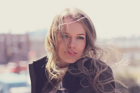 Backlit outdoor portrait of a blonde woman with loose hair and a dark scarf; bright haze and direct gaze create a soft, contemporary editorial mood.