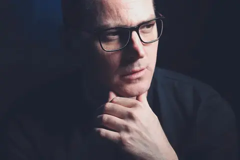 Close executive portrait of a man with glasses resting his hand at his chin; dark background and thoughtful downward gaze communicate analysis, experience and discretion.