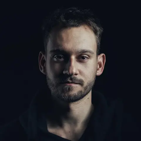 Low-key male headshot against black, with one side of the face emerging from shadow; direct gaze and minimal styling communicate focus, seriousness and modern authority.
